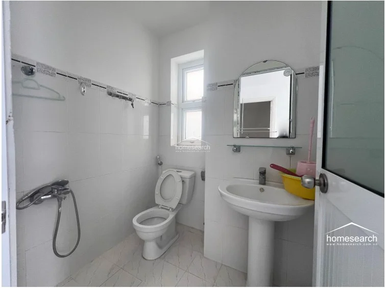 This compact bathroom features white wall tiling with a decorative border and light-colored floor tiles. It includes a pedestal sink, a standard toilet, and a window for natural light, maintaining a clean and functional aesthetic.