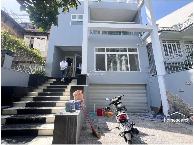 This modern multi-story townhouse features a clean, minimalist grey facade with a prominent front staircase leading to the main entrance. The ground level includes a secure rolling garage door and a paved driveway, while the upper levels offer balconies with glass railings and large windows for natural light.