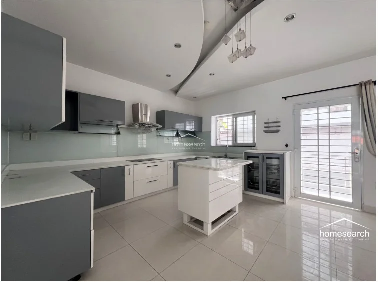 This modern, spacious kitchen features a sleek two-tone grey and white cabinetry design with a central island that provides additional workspace. The room is finished with light-colored floor tiles and a contemporary recessed ceiling with pendant lighting, while a glass-paned door offers natural light and access to an outdoor area.