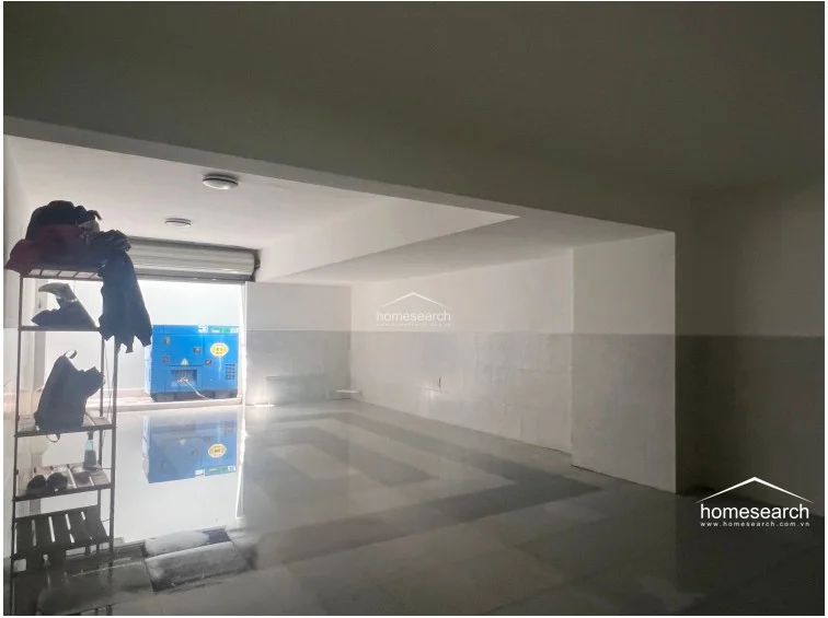This large, open-plan utility or storage space features neutral white walls and polished light-colored flooring, maintaining a clean and minimalist aesthetic. The area includes a roll-up security door and a large blue industrial generator, suggesting it may serve as a garage or a flexible multi-purpose area.
