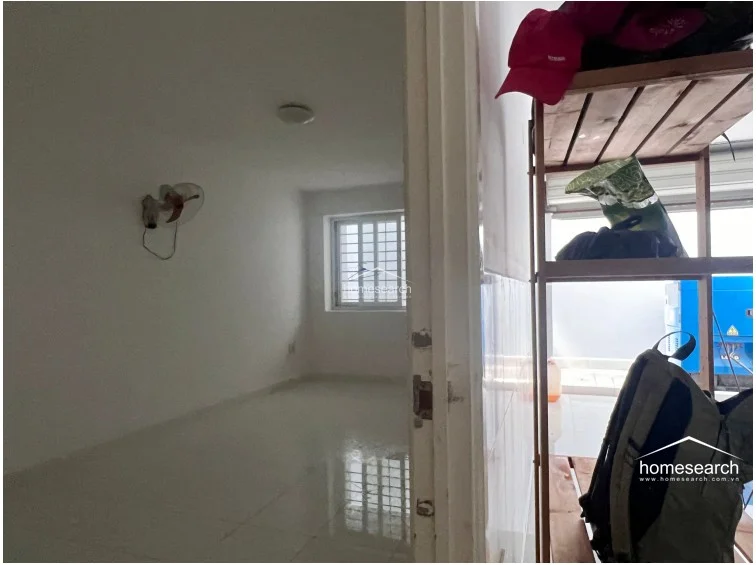 This simple, functional room serves as a bedroom or private study, characterized by its clean white walls and light-colored tile flooring. The space is equipped with a wall-mounted fan and a window that provides natural light, offering a quiet and well-maintained environment.