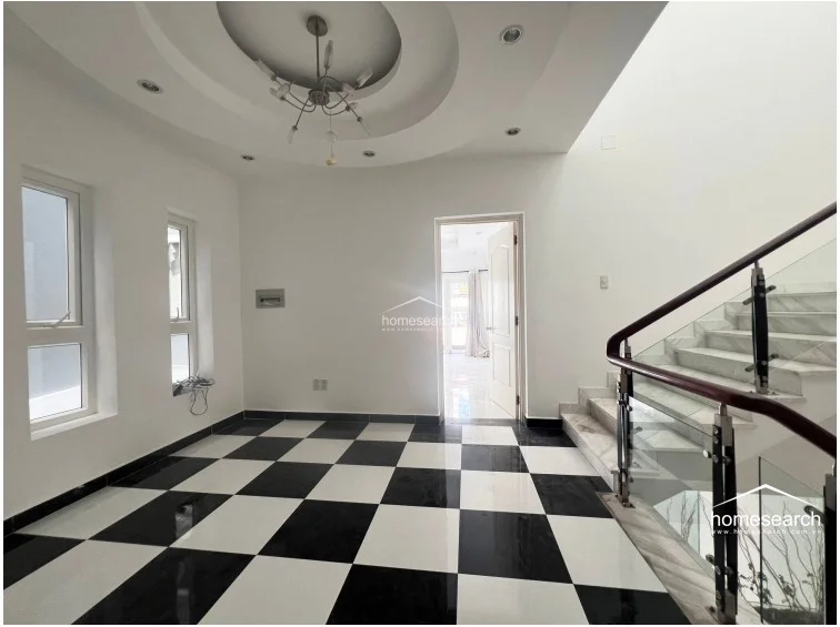 This striking landing or foyer area features a bold black-and-white checkered tile floor and a sophisticated circular recessed ceiling with an elegant modern chandelier. A glass-railed staircase with a dark wood handrail connects the levels, while tall windows ensure the space remains bright and airy.