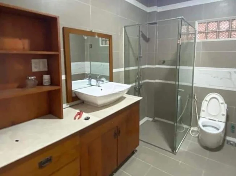 This bathroom features a functional vanity with a wooden cabinet and an open shelving unit, paired with a modern vessel sink. The walk-in shower is enclosed by a glass partition, and the space is finished with neutral-toned wall and floor tiles for a clean, low-maintenance aesthetic.