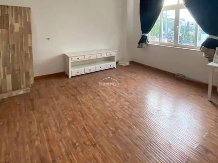 This bedroom features wood-grain flooring and is furnished with a white media console and a matching freestanding wardrobe. The room is accented by dark, heavy curtains that provide privacy and contrast against the neutral walls, maintaining a simple and functional layout.