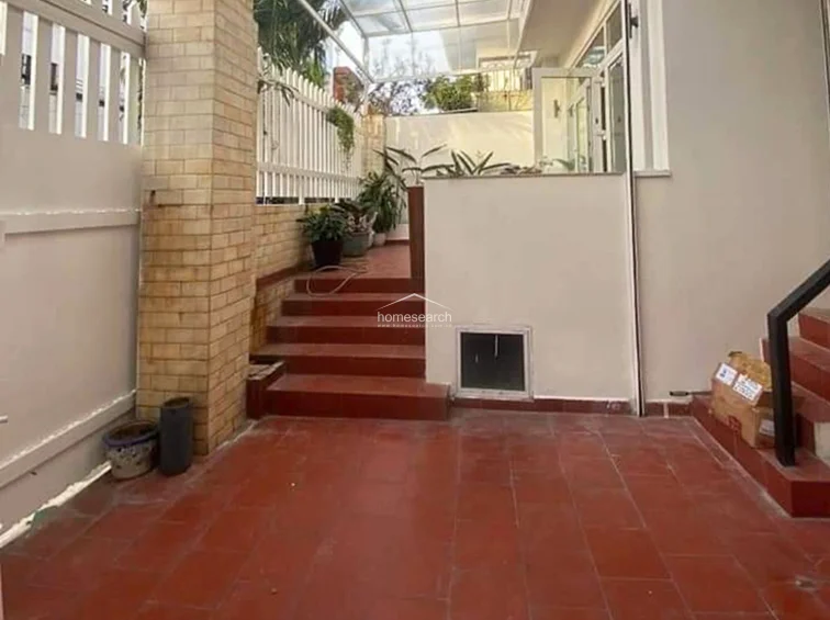 This outdoor terrace features red terracotta-style tiling and a raised platform area with potted plants, providing a clean, multi-level layout. The space is enclosed by a white metal fence and a brick pillar, with a partially covered roof structure that allows for natural light while providing shelter.