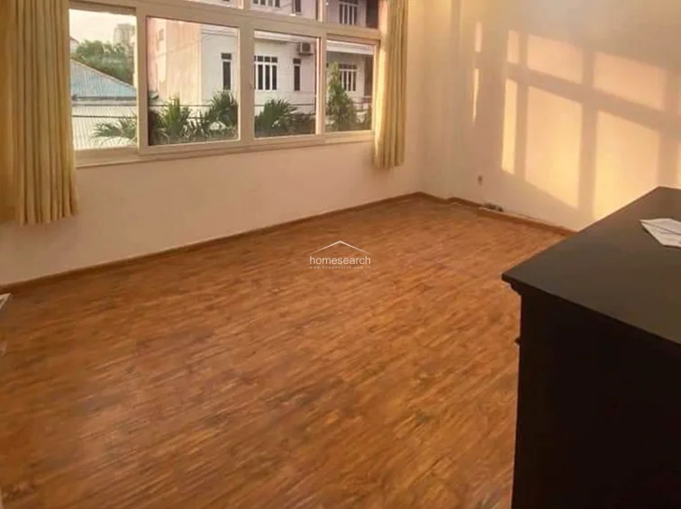 This bedroom or living area is finished with warm-toned wood-look flooring and neutral white walls, creating a bright and inviting atmosphere. Large windows with light curtains allow for significant natural light and offer an exterior view, while the room remains clean and well-maintained.