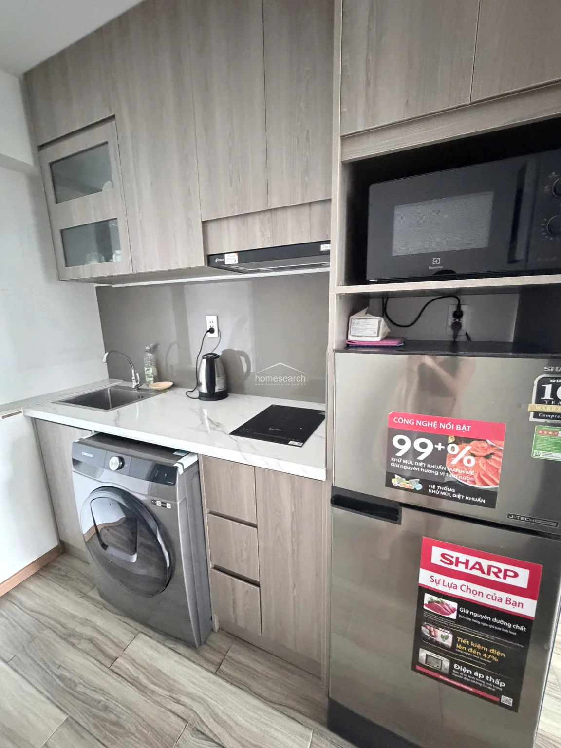 This compact, modern kitchenette features light wood-grain cabinetry and a sleek white marble-look countertop with an integrated induction cooktop and sink. It is highly functional, incorporating a built-in Samsung washing machine, a microwave, and a refrigerator within a space-saving layout.