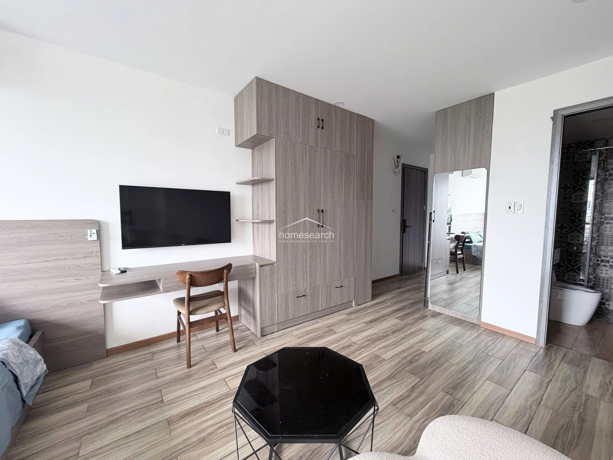 This image showcases the compact yet highly functional kitchenette, which includes a Sharp refrigerator, a built-in induction cooktop, and a Samsung front-load washing machine. The kitchen features a marble-style countertop, a stainless steel sink, and ample overhead cabinetry in a matching wood-grain finish. The space is efficiently organized, making it ideal for urban living with all necessary modern appliances included.