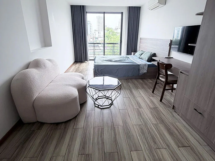 This studio apartment features a modern, open-plan layout with wood-look tile flooring and a neutral white color scheme. The space is anchored by a queen-sized bed and a unique, cloud-shaped sofa, complemented by a wall-mounted TV and a built-in desk area. Large sliding glass doors provide excellent natural light and access to a private balcony with an urban view.