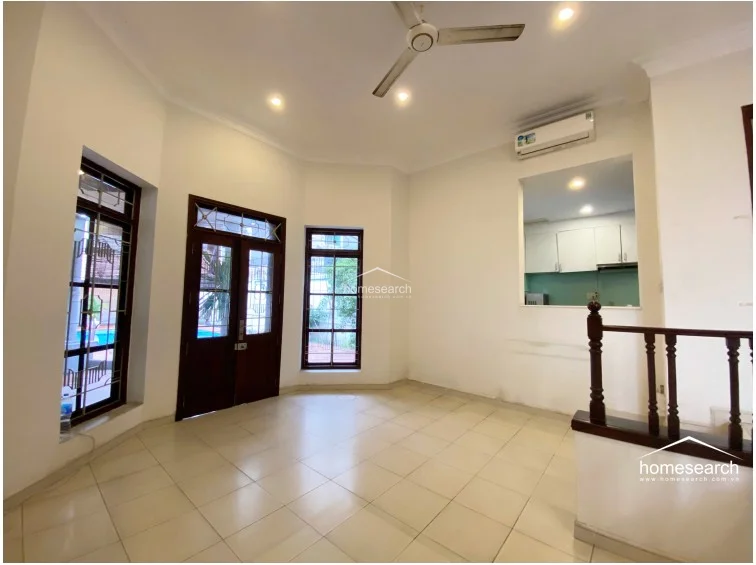 This interior living space features white walls, light-colored tile flooring, and a ceiling fan for climate control. It includes a built-in wall cutout that provides a visual connection to the kitchen area, while a wooden staircase with a traditional balustrade leads to other levels.