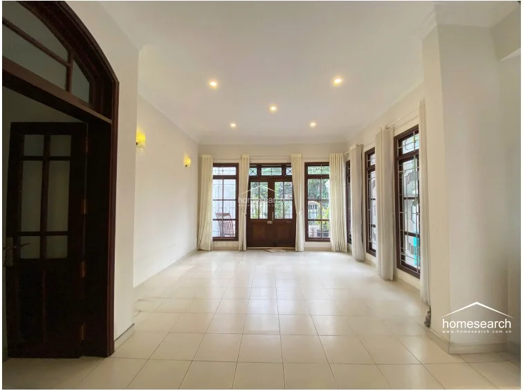 This spacious, open-plan room serves as a central living area, characterized by high ceilings and a clean, minimalist white color scheme. The room is framed by large, dark-wood-trimmed windows and a matching door, creating a seamless visual flow toward the outdoor pool area.