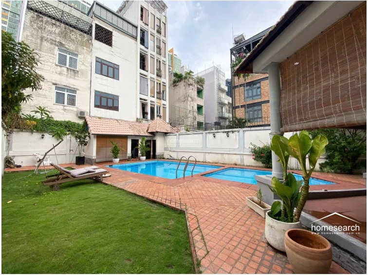This outdoor space features a private swimming pool set within a paved terrace and a small, well-maintained lawn area. The area is enclosed by high perimeter walls for privacy and includes a wooden lounge chair, potted greenery, and a covered patio section with bamboo blinds.