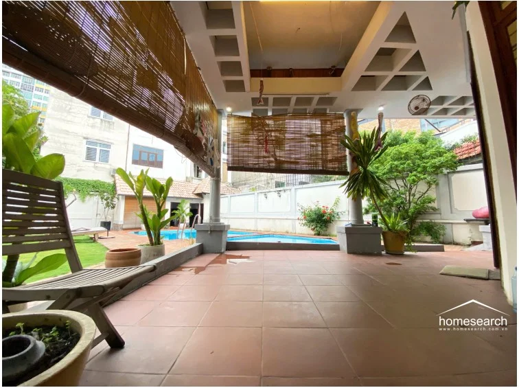 This covered patio area provides a shaded transition between the interior and the pool deck, featuring a unique coffered ceiling design. The space is finished with terracotta-style floor tiles and is equipped with bamboo roller blinds to manage natural light and privacy.