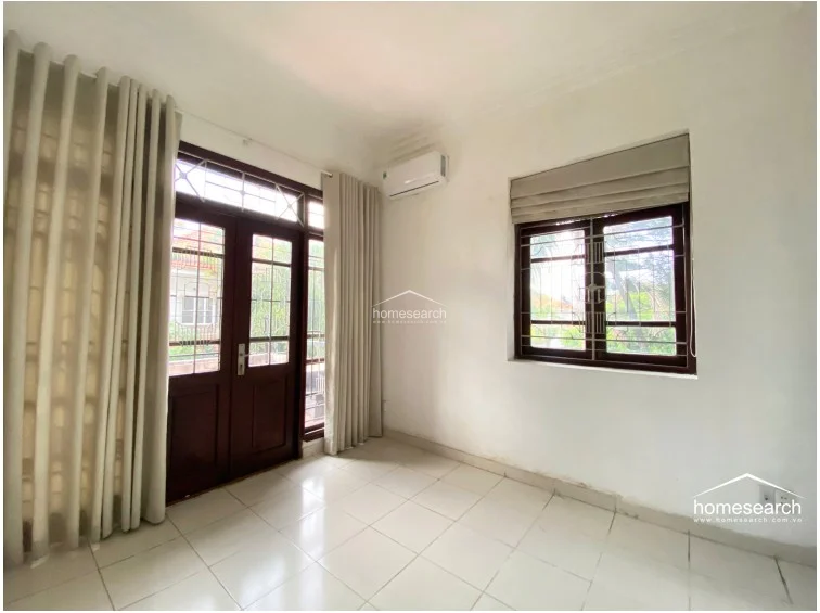 This bedroom features white tiled flooring and neutral walls, creating a clean, minimalist aesthetic. It includes a wall-mounted air conditioning unit, a large window with a Roman blind, and a glass-paneled door that opens to an outdoor space, allowing for ample natural light.