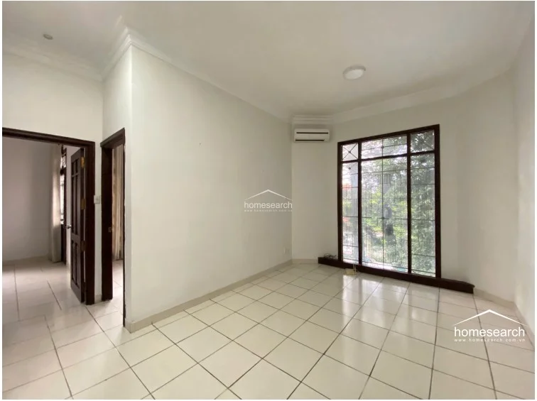 This living or bedroom area showcases a bright, open layout with white tiled floors and white walls, accented by a large window that provides a view of greenery. The space is equipped with a wall-mounted air conditioning unit and features a doorway leading to an adjacent room, suggesting a functional flow between living spaces.