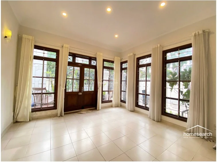 This bright, airy room is enclosed by multiple large windows with dark wood frames and decorative security grilles, allowing for abundant natural light. The space is finished with neutral white walls, light floor tiles, and floor-to-ceiling cream curtains that provide a soft, elegant aesthetic.