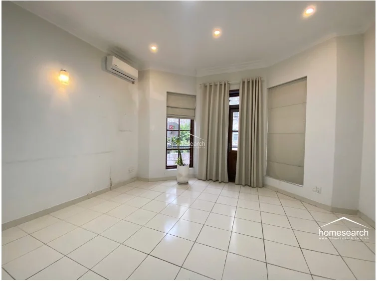 This room is illuminated by recessed ceiling lights and a wall sconce, creating a warm, inviting atmosphere. It features white tiled flooring, a wall-mounted air conditioning unit, and a combination of floor-length curtains and Roman blinds on the windows and door, which offer access to an outdoor area.