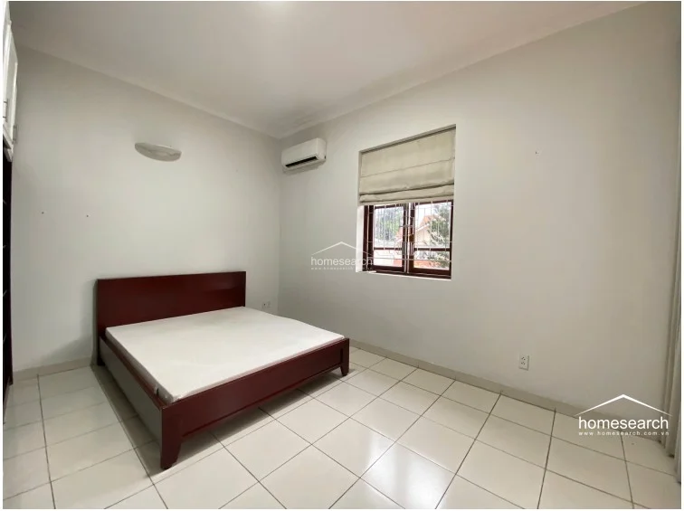 This bedroom is furnished with a dark wood bed frame and features white tiled flooring and walls, maintaining a consistent, neutral design throughout the home. The room is equipped with a wall-mounted air conditioning unit, a window with a Roman blind, and a built-in wardrobe visible on the left, providing practical storage solutions.