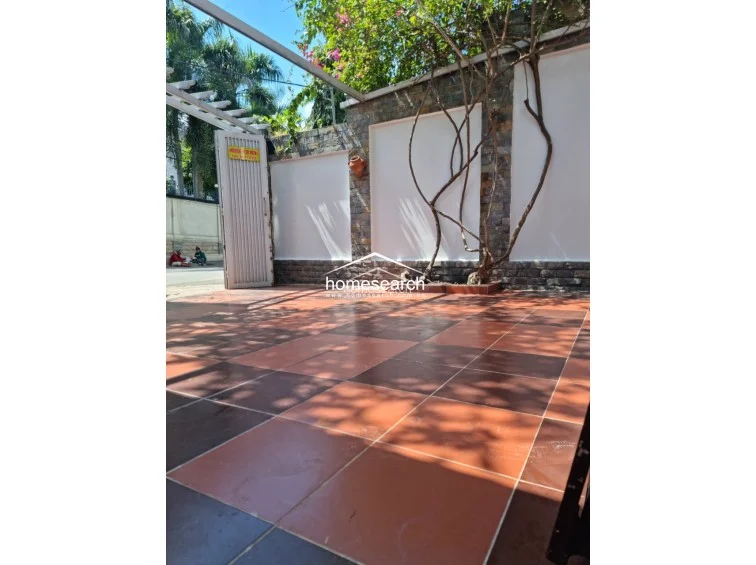 This image shows a private courtyard or driveway area paved with a two-tone terracotta tile pattern. The space is enclosed by white walls accented with natural stone pillars and features a pergola structure, offering a well-maintained, semi-outdoor transition space.