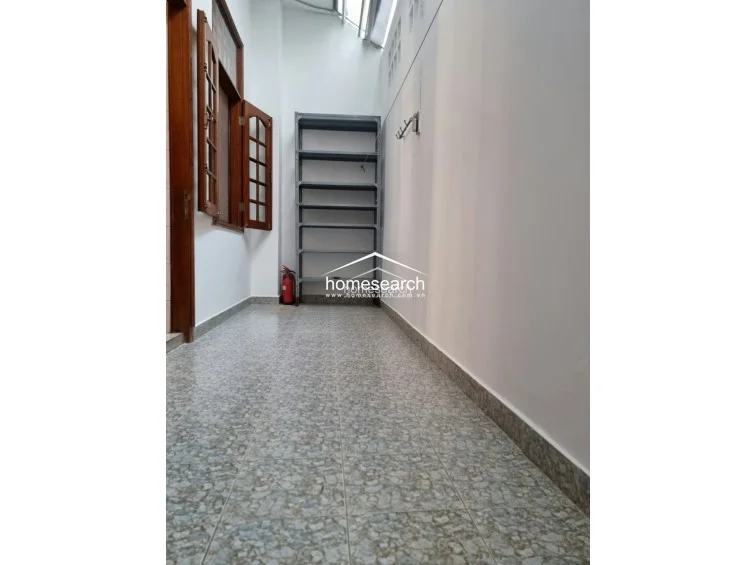 This narrow, covered side passage or utility area features light-colored speckled tile flooring and white walls. It includes a tall metal shelving unit for storage and provides access to the interior via wooden-framed glass doors.