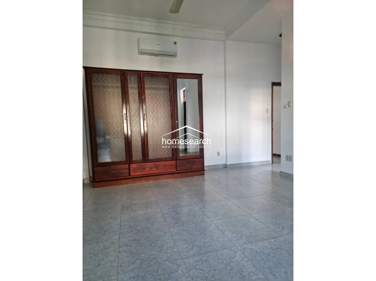 This bedroom or living space features neutral white walls, light grey speckled tile flooring, and a large, dark wood wardrobe with glass panels. The room is equipped with a wall-mounted AC unit and a ceiling fan, maintaining a clean and functional aesthetic.
