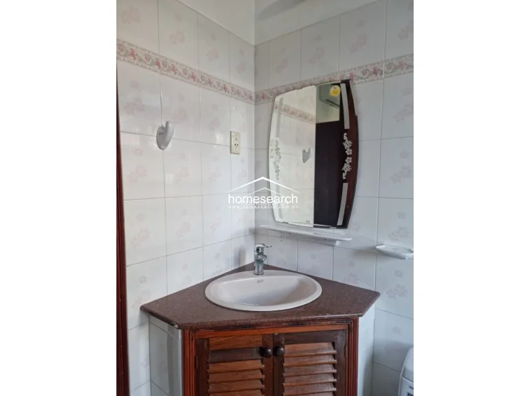 This bathroom features a space-saving corner vanity with a dark wood cabinet and a granite countertop. The walls are finished in light-colored ceramic tiles with a decorative floral border, and the space includes a uniquely shaped mirror with floral glass etchings.
