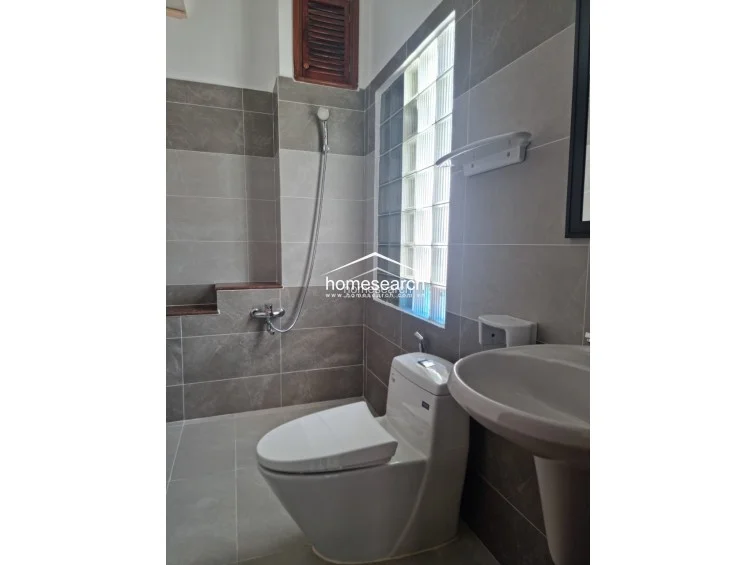 This modern bathroom is finished with neutral-toned, large-format wall and floor tiles, creating a sleek and cohesive aesthetic. It is equipped with a contemporary white pedestal sink, a wall-mounted toilet, and a glass block window that provides privacy while allowing ample natural light to filter in.