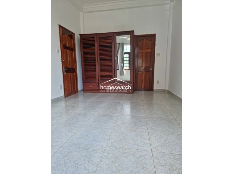 This bedroom features light-colored, patterned tile flooring and crisp white walls, providing a bright and neutral backdrop. A substantial, custom-built wooden wardrobe with louvered doors and an integrated mirror serves as the focal point, offering excellent storage capacity.