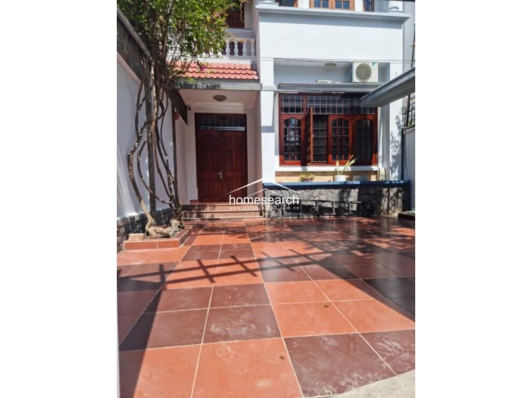 This exterior view shows a paved terrace area featuring a mix of red and brown square tiles, leading to a wooden front door. The space includes a climbing plant feature, a stone-accented wall, and large windows with wooden frames, suggesting a well-maintained, traditional-style entrance.