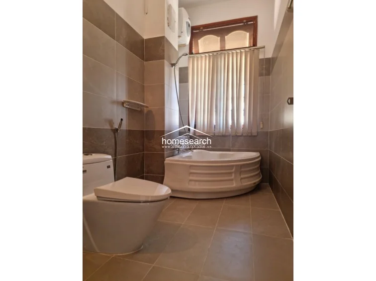 This bathroom is finished in neutral-toned wall and floor tiles, featuring a corner soaking tub positioned beneath a window covered by vertical blinds. The space includes a modern white toilet and a handheld shower fixture, presenting a clean and functional design.