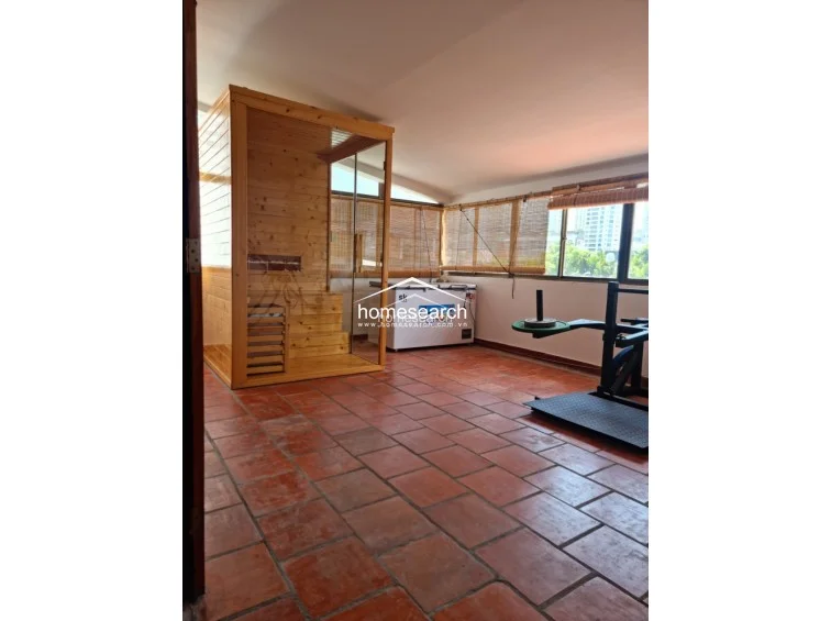 This versatile space is currently utilized as a home gym, featuring durable red terracotta-style tile flooring and a built-in wooden sauna unit. Large windows with bamboo blinds allow for natural light, and the room provides ample open space for fitness equipment.