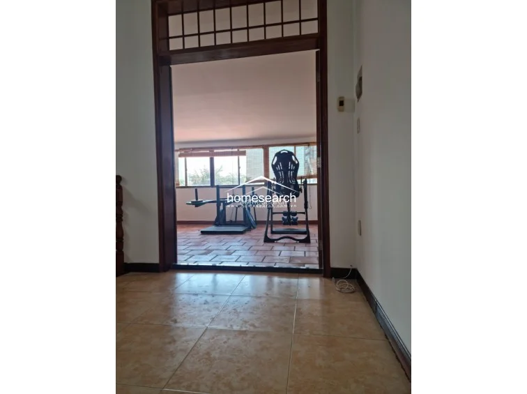 This view captures a transition from an indoor tiled hallway into a covered outdoor terrace or balcony area featuring terracotta-style flooring. The space is currently utilized as a home gym with fitness equipment and provides ample natural light through a large window wall.