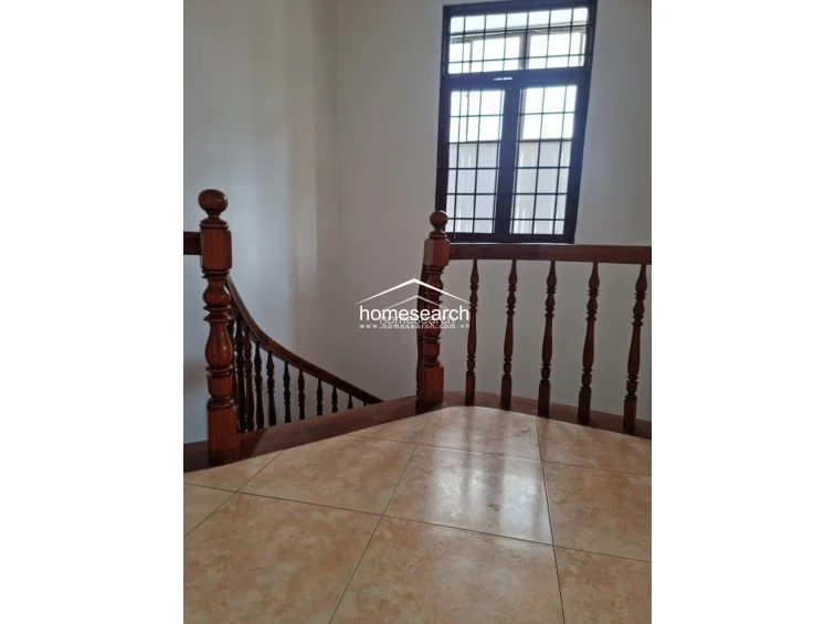 This perspective highlights a staircase landing area featuring a classic wooden balustrade and banister. The flooring transitions to a warm, earth-toned tile, and a large window with a grid pattern provides natural illumination to the stairwell.
