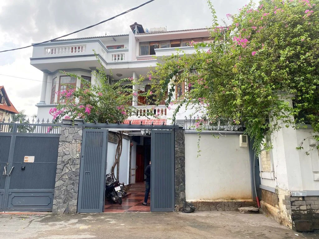 The exterior facade of the property features a multi-level design with white walls, stone-clad pillars, and dark-framed wooden windows. A metal gate provides security, while mature bougainvillea adds a vibrant, welcoming touch to the front entrance.