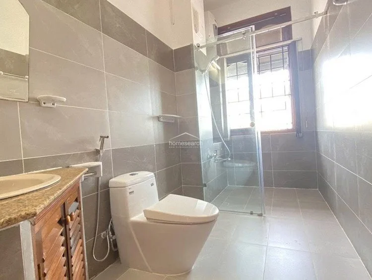 This bathroom features a contemporary design with dual-tone grey wall tiles and light-colored floor tiles. It is equipped with a glass-enclosed shower stall, a modern toilet, and a wooden vanity with a stone countertop, all appearing to be in good, well-maintained condition.