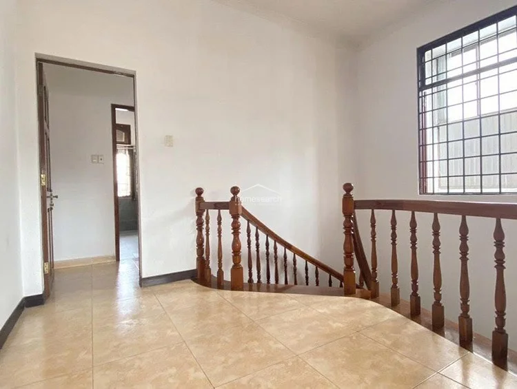 This landing area serves as a central circulation point, connecting to other rooms via wooden doors and featuring a large window secured with a metal grill. The flooring consists of light, neutral tiles that contrast with the rich, dark wood of the staircase railing, maintaining a clean and well-maintained appearance.