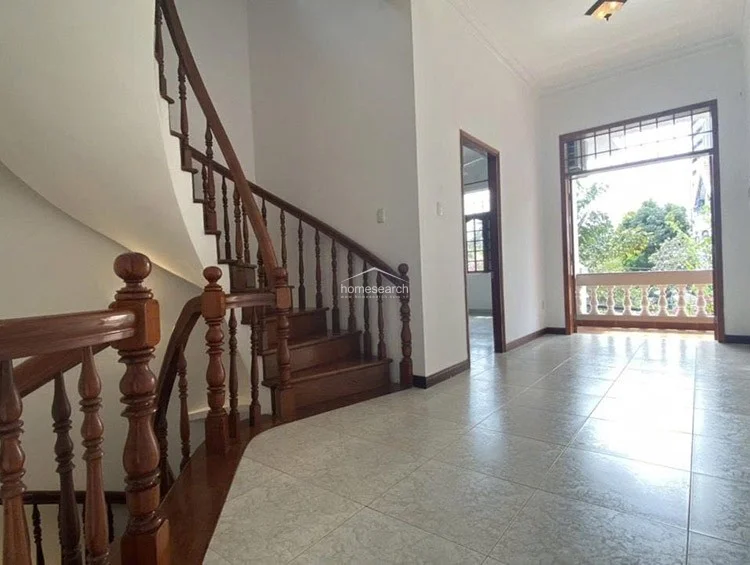 This image shows a bright, open landing area featuring a classic curved wooden staircase with ornate balusters. The space is finished with light-colored floor tiles and white walls, leading out to a balcony with a decorative stone railing that offers an airy, outdoor connection.