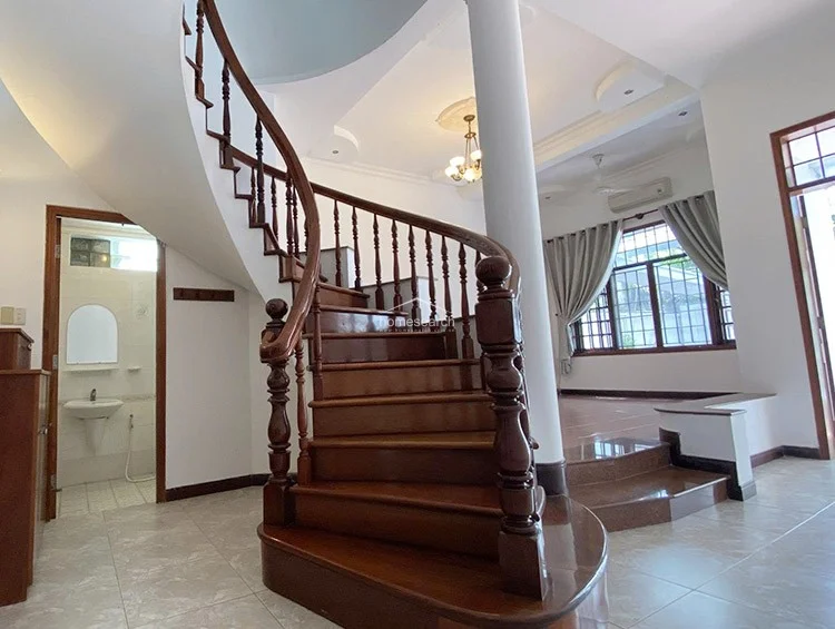 This grand entrance area showcases a striking curved wooden staircase with ornate balusters, serving as the focal point of the home. The open-plan layout connects to a sunken living area and a ground-floor bathroom, all finished with polished light-toned floor tiles and high ceilings.