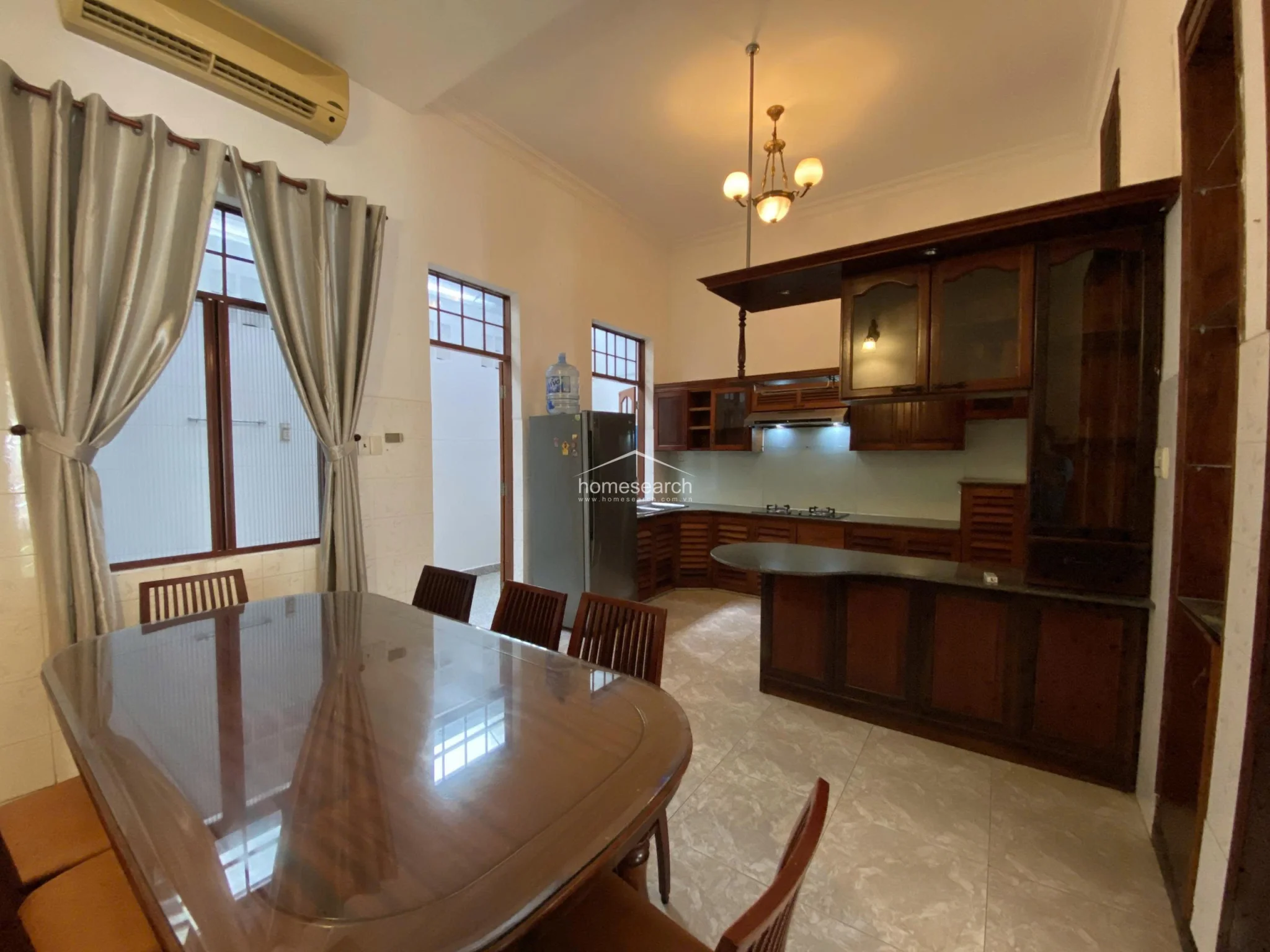 This open-concept kitchen and dining area features a large wooden dining set and a U-shaped kitchen with dark wood cabinetry and a breakfast bar. The space is well-lit by a decorative chandelier and natural light from the windows, complemented by light-colored floor tiles and a wall-mounted air conditioning unit.