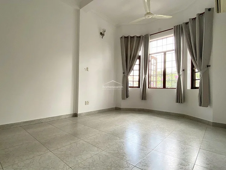 This bright bedroom features a curved bay window layout that maximizes natural light and offers a pleasant exterior view. The room is finished with neutral white walls, a ceiling fan, and light-colored tile flooring, providing a clean and minimalist aesthetic.