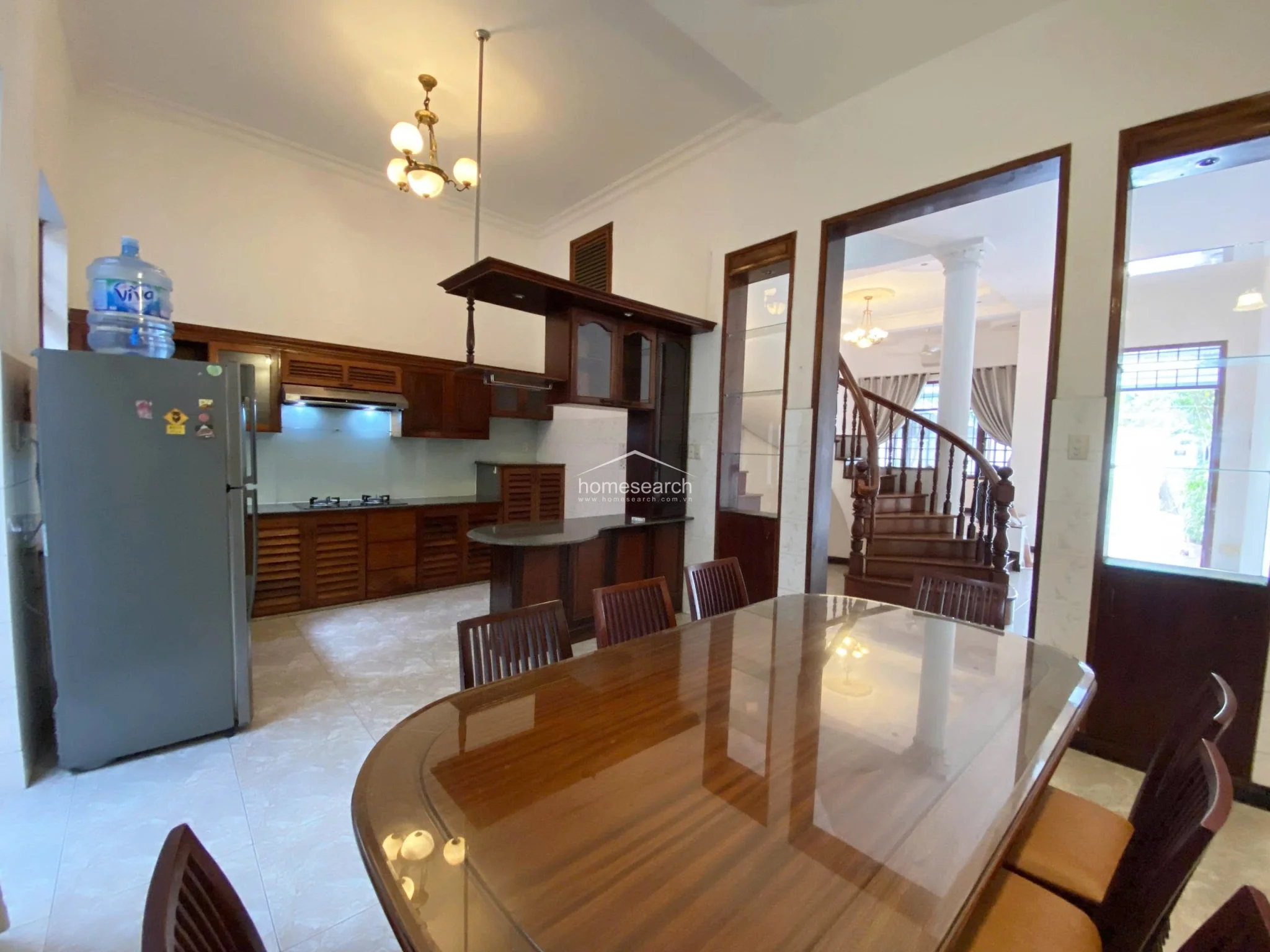 This spacious open-plan area features a large, polished wooden dining table and a well-appointed kitchen with dark wood cabinetry and a breakfast bar. The space is connected to a grand staircase with an elegant spiral design and classic columns, showcasing a sophisticated architectural layout with high ceilings and warm lighting.