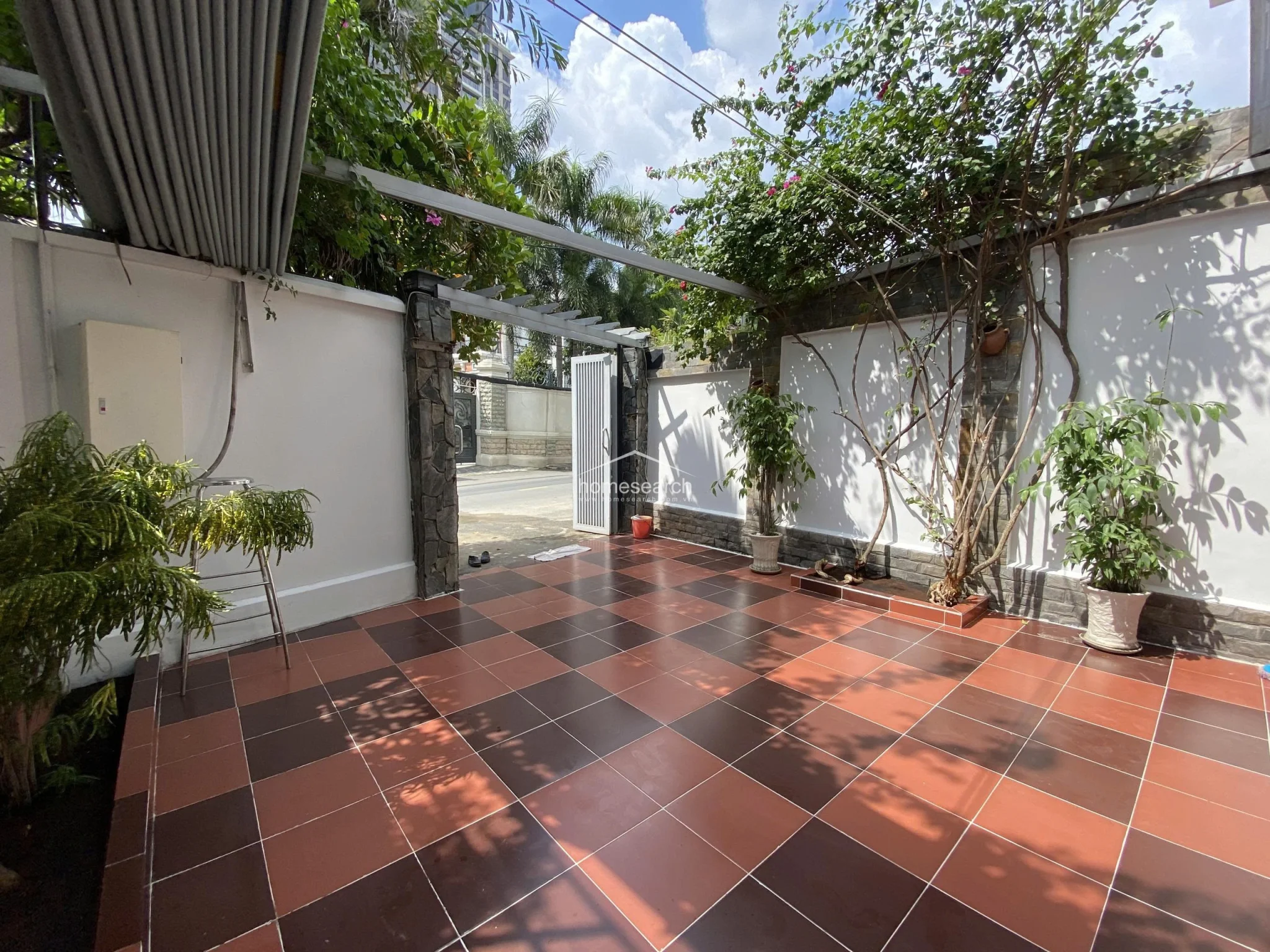 This outdoor courtyard features a checkerboard-patterned tile floor and is framed by white walls with stone-accented columns. The space is partially shaded by a metal pergola and lush climbing vines, offering a private, low-maintenance area for outdoor relaxation.