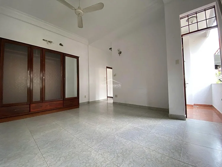 This bedroom includes a large, freestanding dark wood wardrobe with integrated mirror panels, offering significant storage capacity. The room connects directly to a private balcony through a glass-paned door and features light-toned tiled flooring throughout.