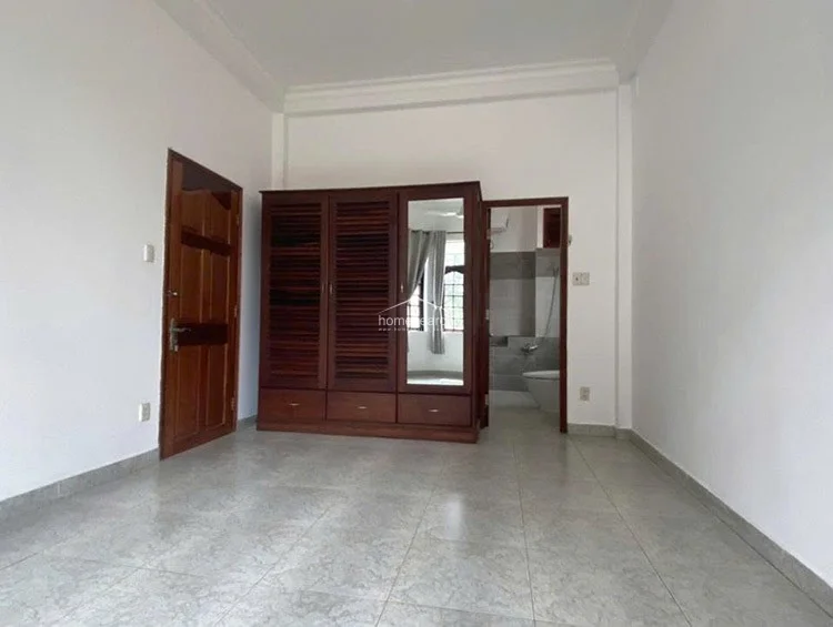 This bedroom layout centers around a large, dark-wood louvered wardrobe that provides both storage and a classic aesthetic contrast to the white walls. The room flows directly into an en-suite bathroom, with light-colored tile flooring and a solid wood entry door completing the functional design.