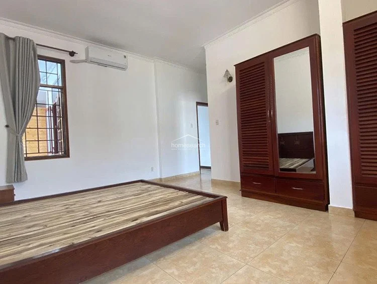 This bedroom view highlights a spacious layout with a dark wood bed frame, a tall matching wardrobe with a central mirror, and an AC unit mounted above the window. The room maintains a clean, neutral aesthetic with white walls, crown molding, and light-colored ceramic tile flooring.