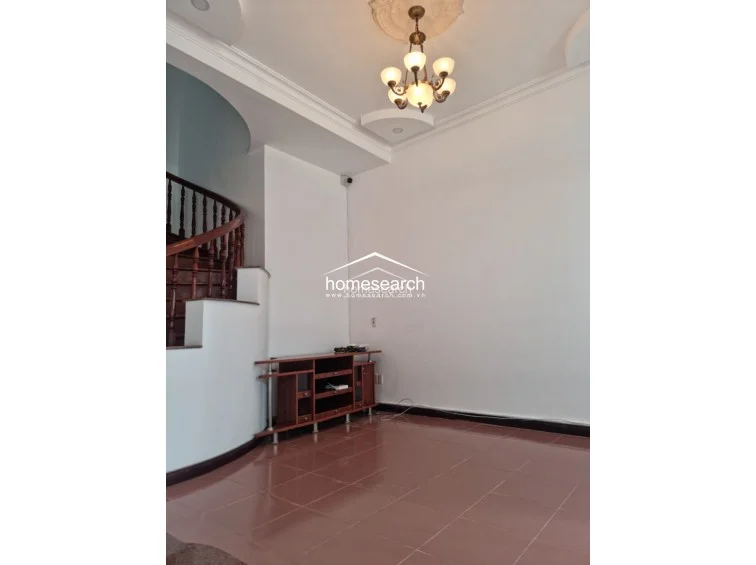 This image shows an open-plan living space characterized by a striking curved wooden staircase with ornate balusters. The room is finished with white walls, decorative ceiling molding, and a traditional chandelier, providing a classic aesthetic that flows into the adjacent areas.