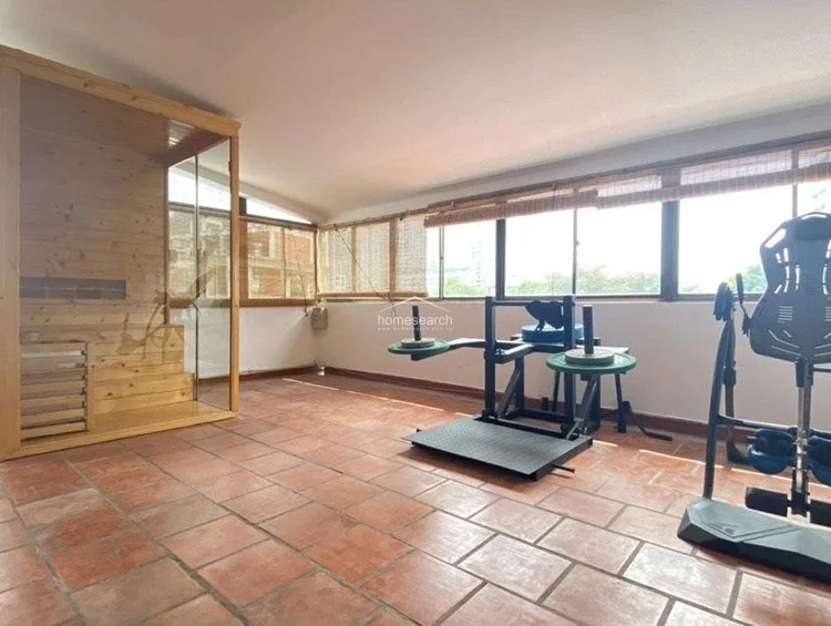 This spacious home gym is finished with rustic terracotta-style floor tiles and features a built-in wooden sauna unit. Large windows with bamboo blinds wrap around the room, providing abundant natural light and a view of the surrounding greenery.