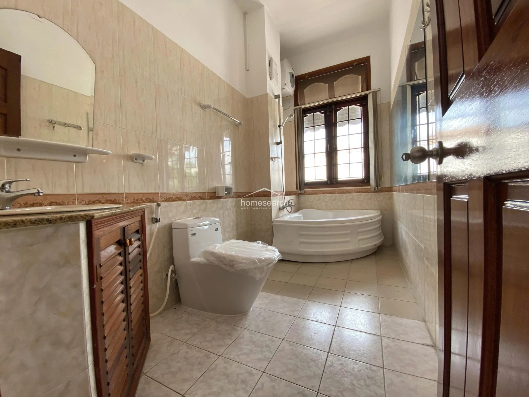 This bathroom features a classic design with light beige wall tiles, a corner soaking tub, and a wooden vanity cabinet with a stone countertop. The room is well-lit by a window with a decorative grill and includes a modern toilet, presenting a clean and functional space.