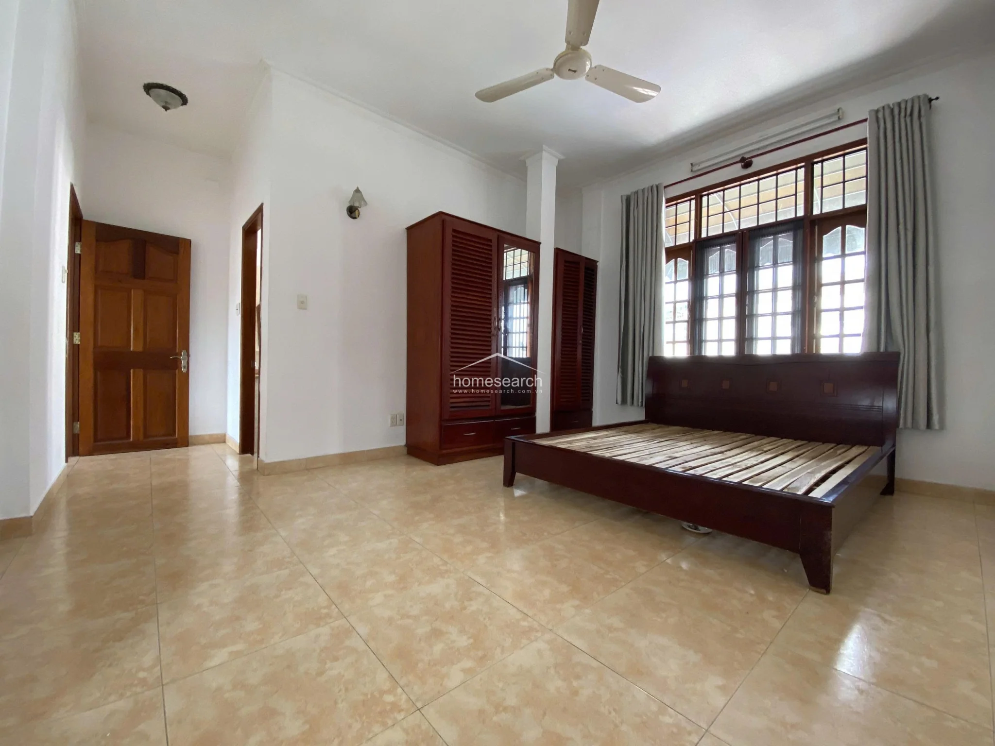 This bedroom interior displays a wide-angle view of the space, showcasing a ceiling fan, a dark wood bed frame, and a large, multi-pane window with grey curtains. The room is finished with white walls and light-colored tile flooring, with a dark wooden door leading to an adjacent area.