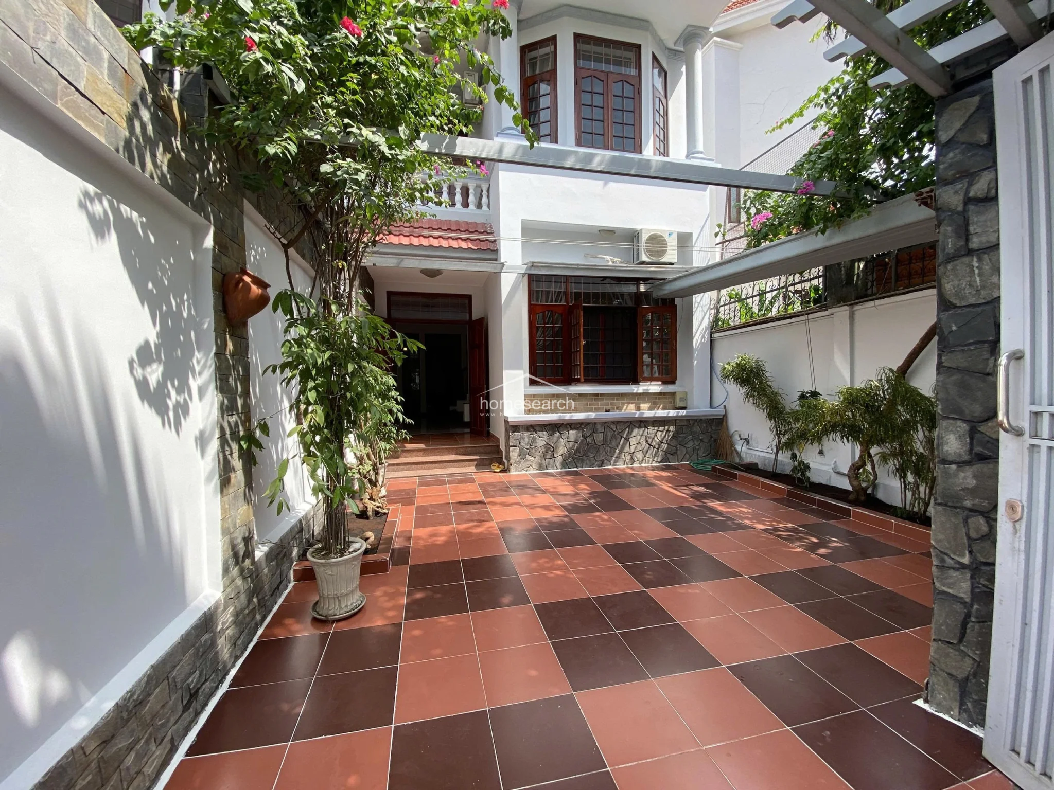 This view highlights the courtyard’s layout, showing how the terracotta tiled ground connects to the main entrance steps and the stone-accented exterior of the house. The space is shaded by a pergola and mature plants, creating a pleasant, private outdoor area that appears to be in good condition.
